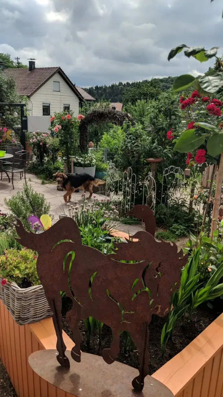 Garten mit Blumen und einer Metallfigur, im Hintergrund Dächer und Hügel