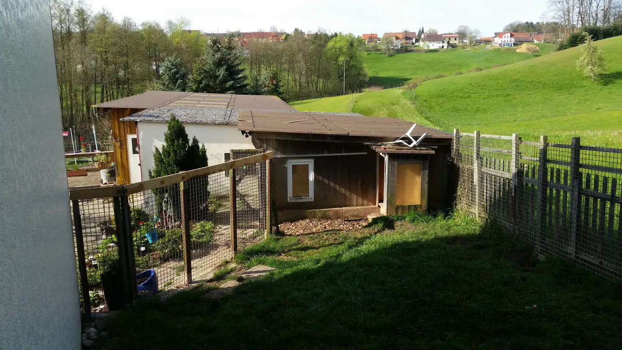 Holzhütte mit Zaun am Hang, dahinter Wiesen und Hügel
